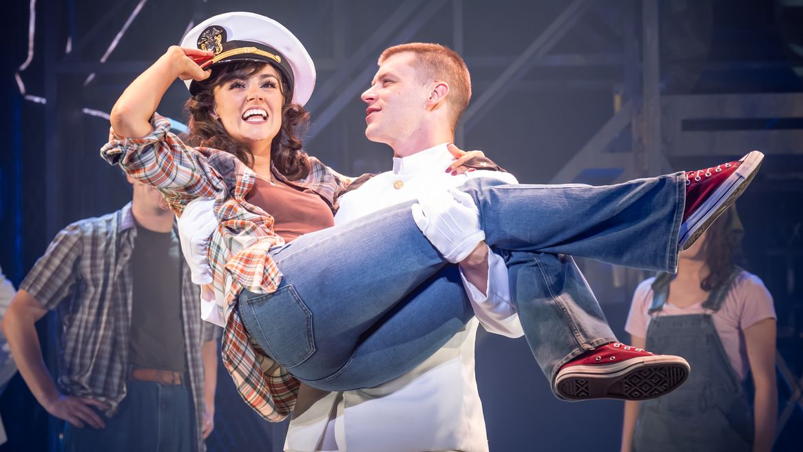 A young man in a white flight officer outfit carries a young woman who is wearing his hat in his arms