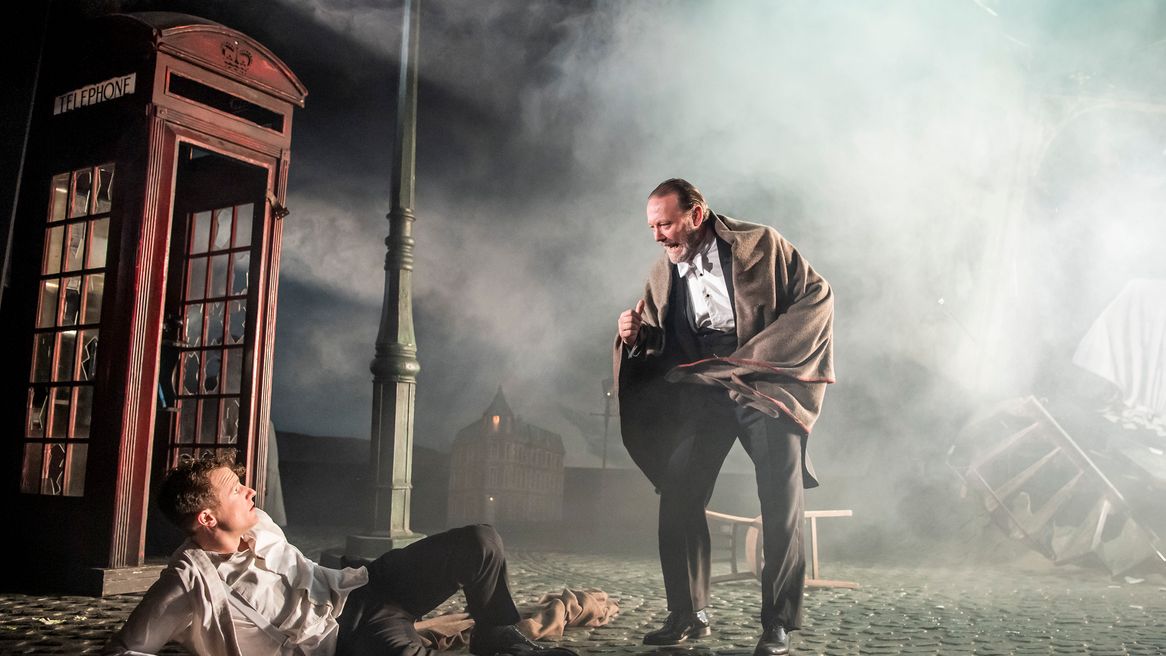 A man lays in a smoke filled cobbled street, while another man in a blanket stands over him with a fist raised. 