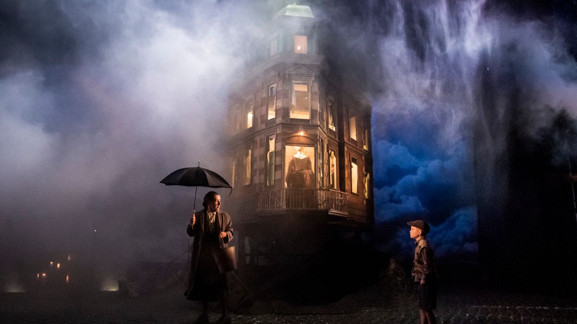 Rain pours down on an illuminated house, in front of which stands a man with an umbrella and a small boy