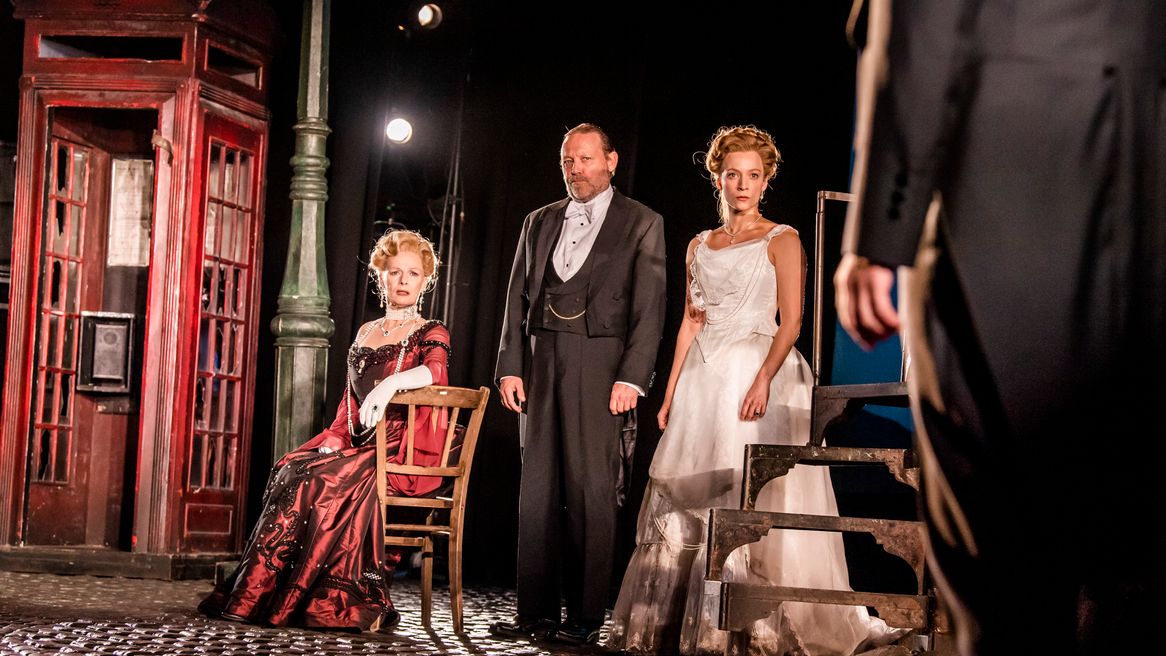 A lady in a red dress sits on a chair next to a man in a dinner suit and another lady in a white dress all look at a figure.