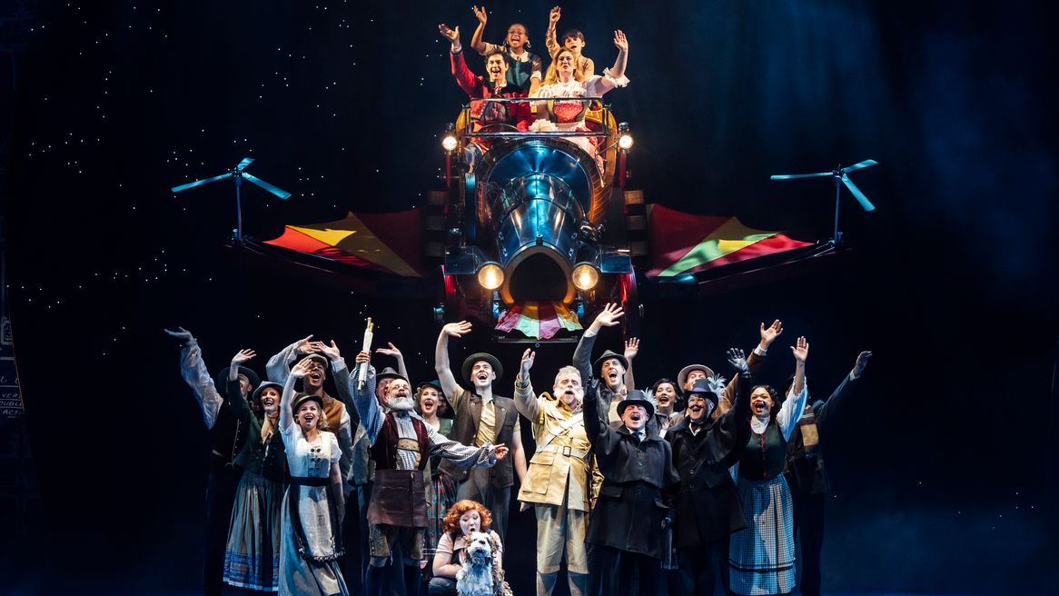 People stand on stage waving at a family in a flying car