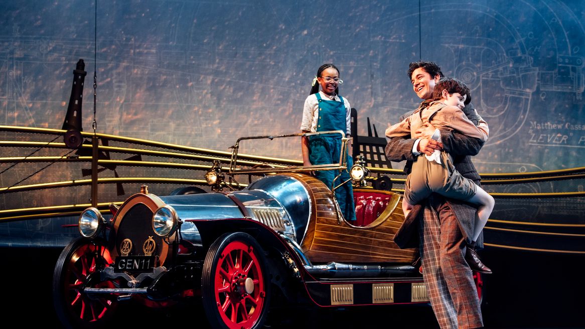 A man hugs his son in front of an old fashioned car while his daughter smiles