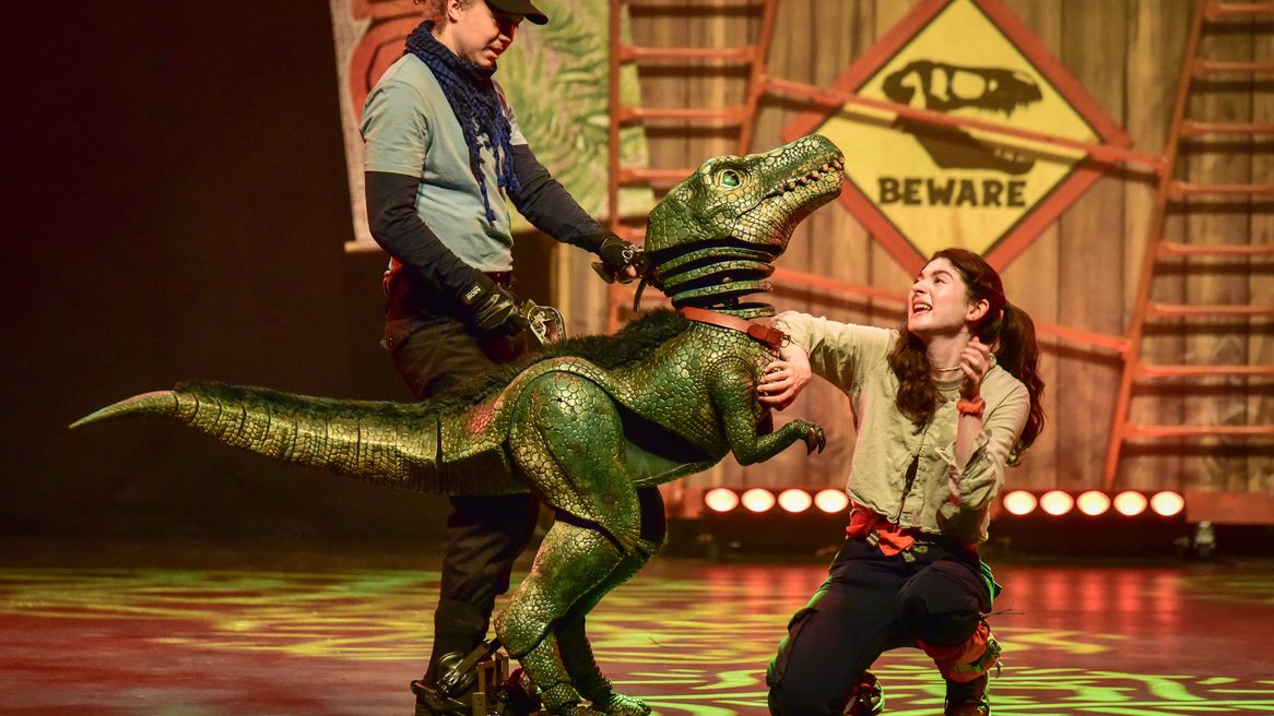 A man controls a small velociraptor puppet while a woman kneels in front