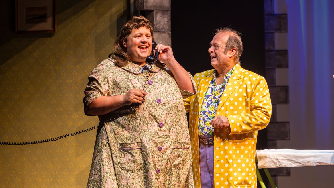 A man dressed as a woman in a plain smock on the telephone with a man in a bright coloured outfit 