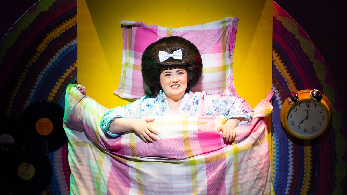 A girl with big hair lies in bed beside a large clock