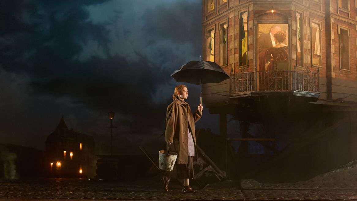 A drably dressed young woman walks past a grand looking house holding an umbrella