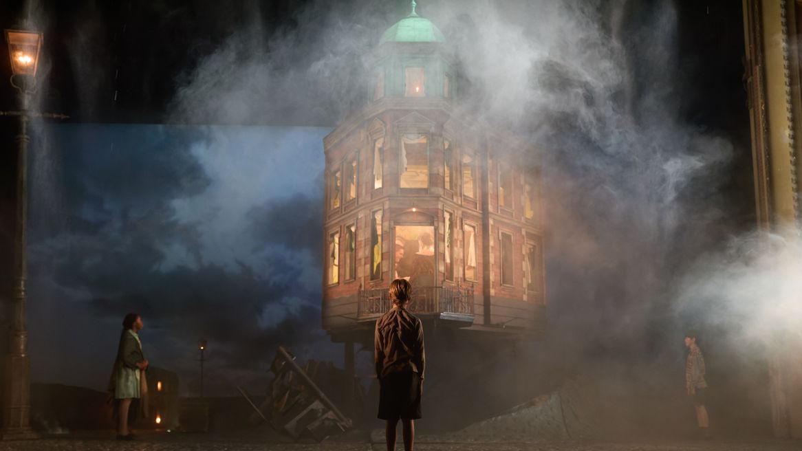 Three poor children stand in smoke covered rubble staring up at a grand house