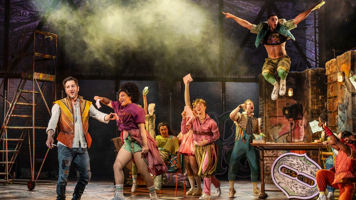 mixed group of dancers waving pieces of paper in their hands while one man leaps from a table in the background.