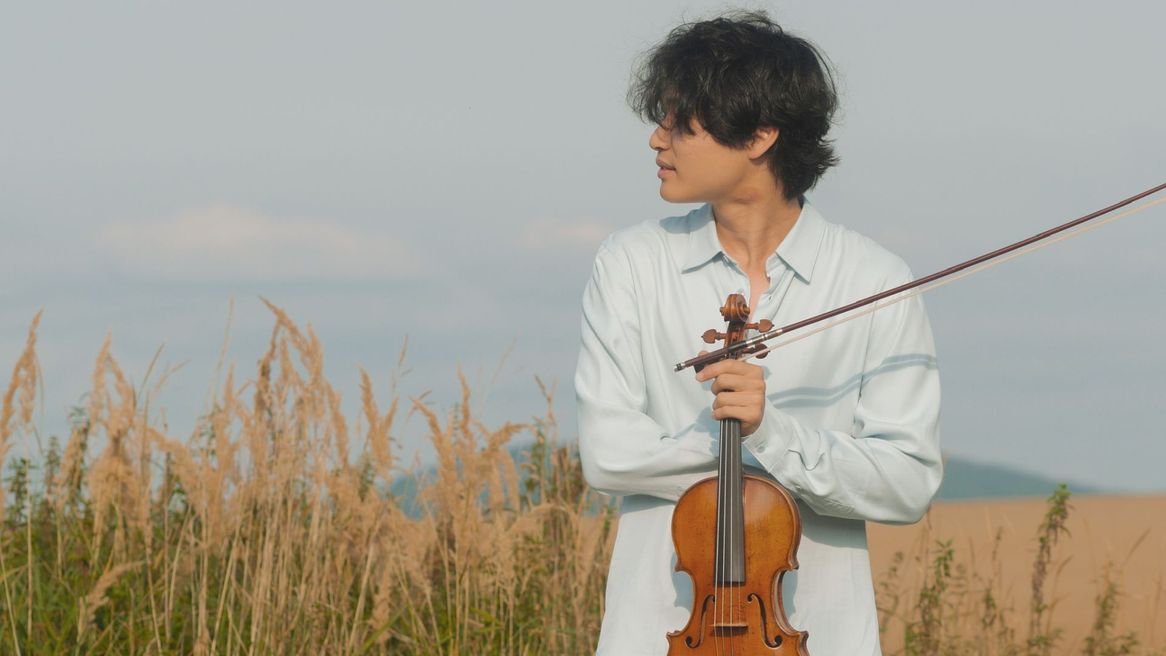 A young man stands in a field holding a violin