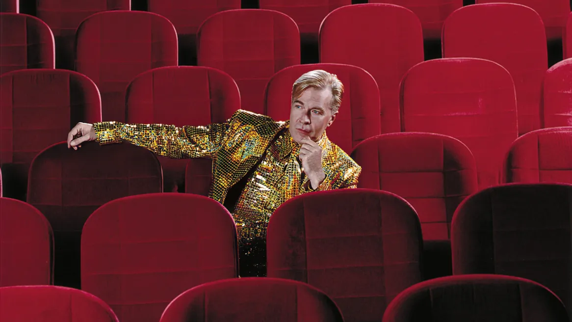 A man in a sequinned gold suit sits alone in an empty theatre