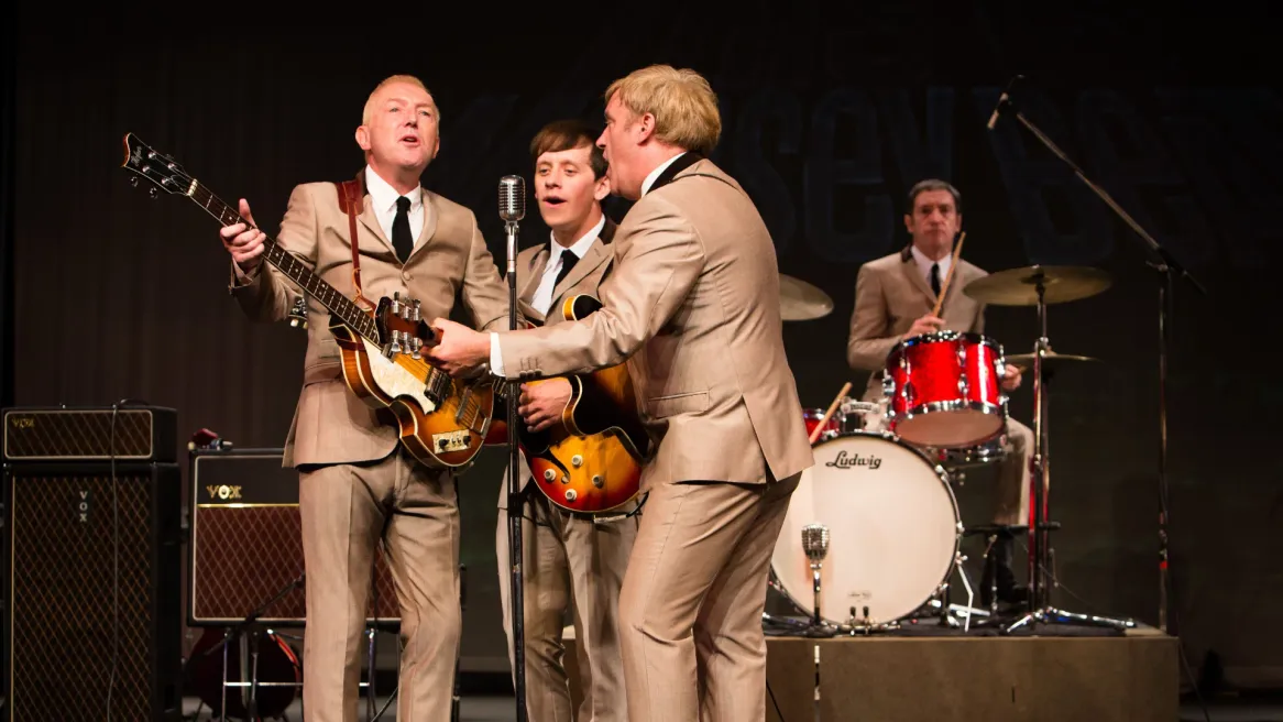 A group of men in grey suits play guitars with a drummer in the background