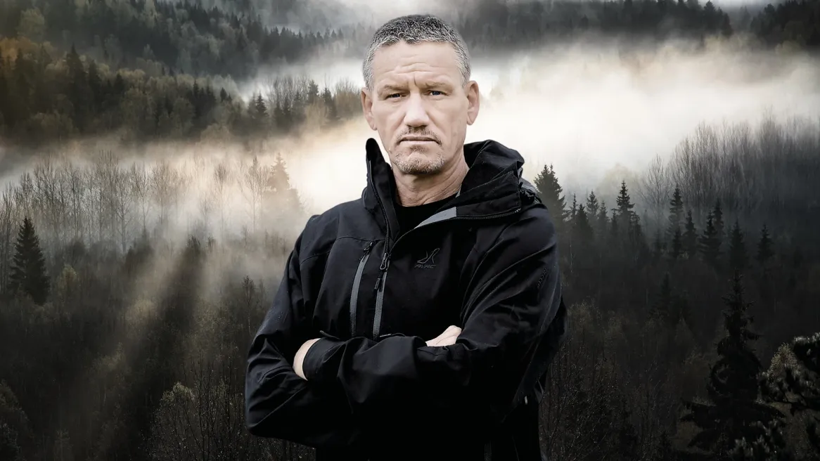 A man dressed in black hiking gear crosses his arms in front of a forest