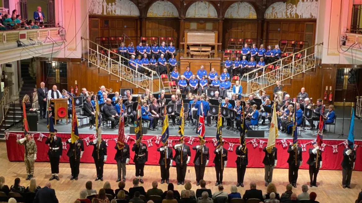 Military members stand in front of a choir and orchestra on stage holding various flags